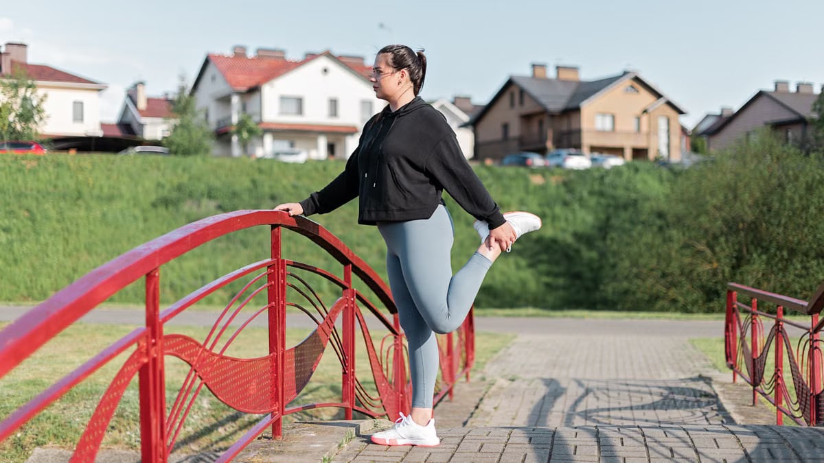 A woman in active wear performs a stretching exercise on a red bridge in a suburban park setting.